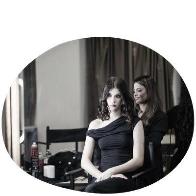 A woman in a black off-shoulder dress sits in a makeup chair, while another stylist works on her hair in a softly lit room.